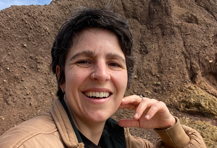 A headshot of Ryat Yezbick. They are seated in front of large brown rock formation.